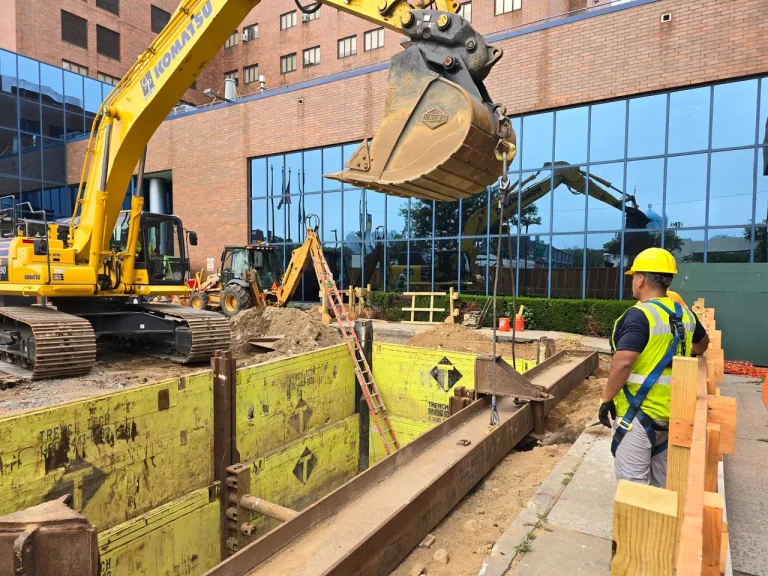 A worker in a hard hat and safety vest beside an excavator on a city jobsite